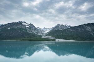 Beautiful snowy mountain peaks rising up from the shoreline of a calm body of water with a stunning reflection below and a cloudy sky overhead. photo