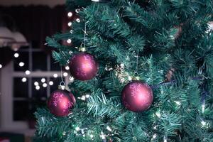 3 red Christmas tree balls hanging by a hook on a branch of an artificial pine tree decorated with lights. photo