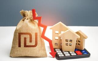 Israeli shekel money bag with down arrow and houses on calculator. Reducing maintaining cost, increasing energy efficiency. Falling real estate market, low prices and demand. Saving resources. photo