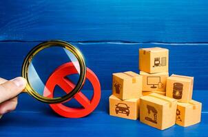 Man with a magnifying glass examines red prohibition sign NO near boxes. Assessment of damage from sanctions or embargo. Effect of restrictive measures. Protect national producers from foreign goods photo