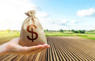 A hand holds out a dollar money bag on a background of a farm field. Lending farmers and agricultural enterprises for purchase land and seed material, equipment modernization Support and subsidies. photo
