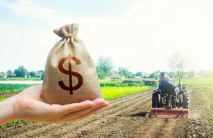 A hand holds out a dollar money bag on a background of a field cultivating tractor. Lending farmers for purchase land and seed material, modernization. Support and subsidies. photo