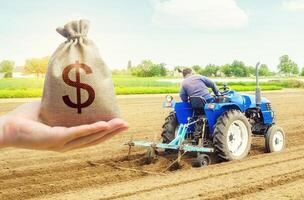 A hand holds out a dollar money bag on a background of farmer on a tractor making mounds rows on a farm field. Lending farmers for purchase land and seed, modernization. Support subsidies. Farm loans photo