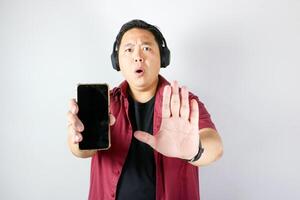 Handsome man showing rejection hand gesture and saying NO pose while wearing headphone and showing his blank screen smartphone over isolated white background. photo