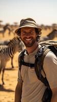 Man wearing a safari hat and backpack smiling with zebra in the background photo