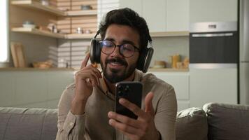 Relaxed happy man Indian Arabian guy male smiling choose audio song playlist on mobile phone smartphone listening music in wireless headphones earphones enjoy sound lazy weekend relax on couch at home photo