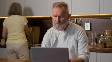 Middle aged retired 50s man husband gray-haired businessman thinking working distant with computer notebook thoughtful think problem solution laptop work at home while woman wife cooking in kitchen photo