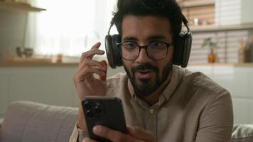 Happy smiling Indian man contented Arabian guy listen music in headphones enjoy song relax at home kitchen listening sound on mobile phone musical playlist online audio service in wireless earphones photo
