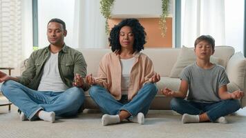 Calm peaceful multiracial family African American parents teach meditation son kid with closed eyes do yoga exercise mom dad and child little boy relax together in lotus pose zen balance on home floor photo
