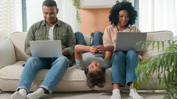 Addicted family use gadgets devices little kid child son boy lying upside down on sofa play game on mobile phone smiling African American parents working laptops different gen using modern tech photo