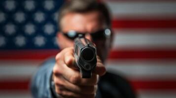 Person aiming a handgun towards the viewer with an American flag backdrop during a dramatic scene in a dimly lit environment photo