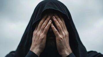Individual in dark hood covering face with hands in an outdoor setting under cloudy sky photo