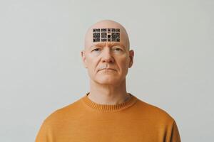 Digital slavery, digital concentration camp, social problem of the future concept. Man with QR codes on forehead poses in a neutral setting while wearing an orange sweater photo