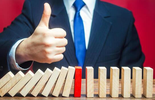 A businessman shows a thumbs up approval over a stopped process falling domino. Find a way stop the problem and prevent its spread. Achieving success and completing a task. Solution. Cyber protection photo