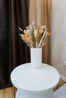 A stunningly crafted vase filled with an assortment of dried flowers in a range of vibrant colors is gracefully positioned on a sleek white table, enhancing the charming decor of the room photo
