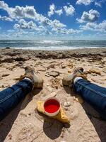 A person is relaxing while laying on the warm beach, holding a cup filled with a vibrant red liquid that catches the sunlight beautifully, enjoying the serene coastal view photo