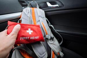A person is standing next to a car while holding a first aid kit in one hand and a backpack resting beside them, all positioned on the ground near the vehicles open door photo