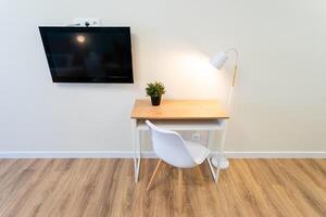 A charming wooden desk with an ergonomic chair and stylish lamp is conveniently placed under a wallmounted flatscreen TV, enhancing the cozy atmosphere of the workspace photo