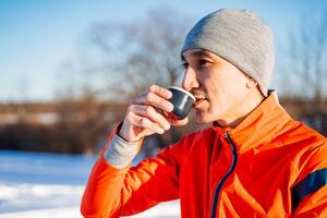 Man drinking tea from mug while standing in nature in winter, man in orange jacket and hat enjoying cup of coffee in winter forest, photo