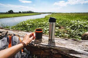 picnic in nature, there is a thermos and a mug on a tree, tourist utensils made of metal, drink hot tea in the forest, summer heat in nature. photo