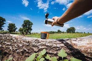 In a peaceful natural setting, someone is pouring water from a thermos into a cup placed on a log, surrounded by trees and greenery. The atmosphere is serene and tranquil photo