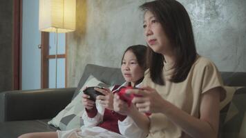 Asian family, mum, and kid fun playing game with joy controllers on sofa in home living room together, enjoy and cheerful, happy weekend exciting hobby of child and parent, digital entertainment. video