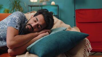 Tired indian man lying down in bed taking a rest at home, napping, falling asleep on sofa, relaxing video