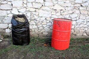 The waste disposal setup includes a black bag and a red barrel placed against a stone wall photo