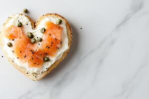A heart-shaped bagel features a generous layer of cream cheese, adorned with slices of smoked salmon and capers, set against a light marble background with plenty of space for text photo