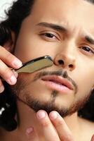 Man with curly hair grooming his beard carefully using a straight razor against a plain white background photo