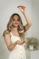 young beautiful blonde girl stands among feathers. feathers fly through the air in a white room. one feather flies between her hands. holds one feather in her hands. one is dressed in a white dress photo