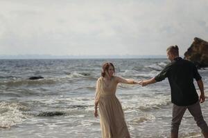 lovers and romantic newlyweds having fun on the seashore. couple dancing on the seashore. they hold hands and look at each other. girl wearing beige dress, wind blowing their hair photo