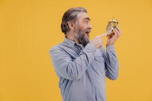 Surprised senior man pointing at alarm clock on yellow background photo