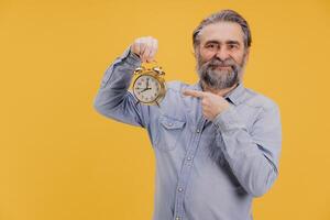 Smiling senior man pointing at alarm clock on yellow background photo