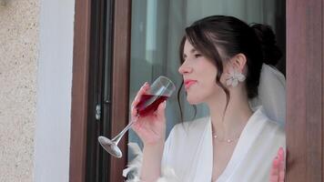 Bride sipping wine in elegant robe, A bride wearing a white robe and floral earrings enjoys a glass of red wine while video