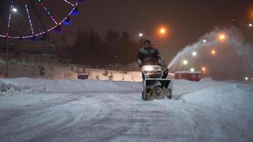 Snow removal with a snow thrower in the evening in an amusement park. A man drives a harvesting manual tractor throwing snow from the track video