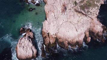 Aerial top view of waves break on rocks in a blue ocean. Stock footage. Sea waves on beautiful beach aerial view drone. Bird's eye view of ocean waves crashing an empty stone rock cliff from above. video