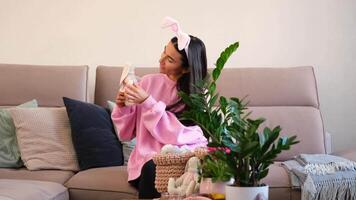 Woman in pink sweater and bunny ears holding a handmade rabbit doll while sitting on a cozy sofa decorated with cushions and plants video