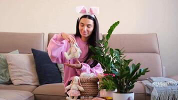 Woman wearing bunny ears holding a stuffed rabbit toy, sitting on a sofa in a cozy living room with a green plant and festive decorations video