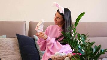 Young woman wearing bunny ears and holding an Easter bunny toy, sitting on a cozy couch with decorative pillows and a green plant in the foreground video
