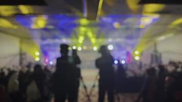 Blurry image showing a crowd of people and stage lighting. Out of focus with yellow and blue lights. Two people can be seen indistinctly in the crowd in the background. video