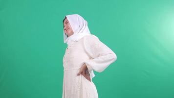 A woman wearing a white dress and hijab shows an expression of pain in her spine while holding it with both hands on a green background or isolated green screen. video