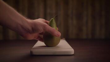 A hand is gracefully holding a ripe pear positioned above a cutting board placed on a table video