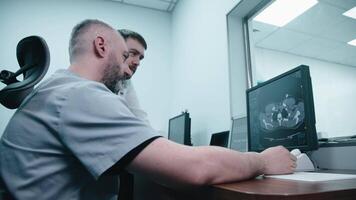 Two men Radiologists Watching Diagnosis Procedure and Monitors on computer Showing Heart Scans Results, In the Background Patient Undergoes MRI or CT Scan Procedure. video