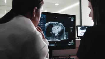 Two Women Doctors, Radiologist Watching Diagnosis Procedure and Monitors on computer Showing Heart Scans Results, In the Background Patient Undergoes MRI or CT Scan Procedure. video