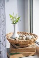 delicate small bouquet of lilies of the valley in a Chinese porcelain vase in the summer on a stack of old books on a windowsill, in a basket with quail eggs, Easter is coming soon, diet breakfast, photo
