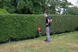 man standing in front of green hedge while working, gardener trimming hedge with electric hedge trimmer,took a break from work photo