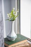 elegant small bouquet of lilies of the valley in a chinese porcelain vase in summer on a stack of old books on a windowsill, photo