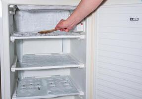 ice has formed on the walls of the freezer and it is creeping out, a woman's hand is knocking the ice off with a wooden spatula, photo