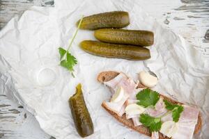 Rustic appetizer of pickles, rye bread with bacon, garlic and parsley on white paper, laid out on a weathered wooden surface with a shot of vodka, photo